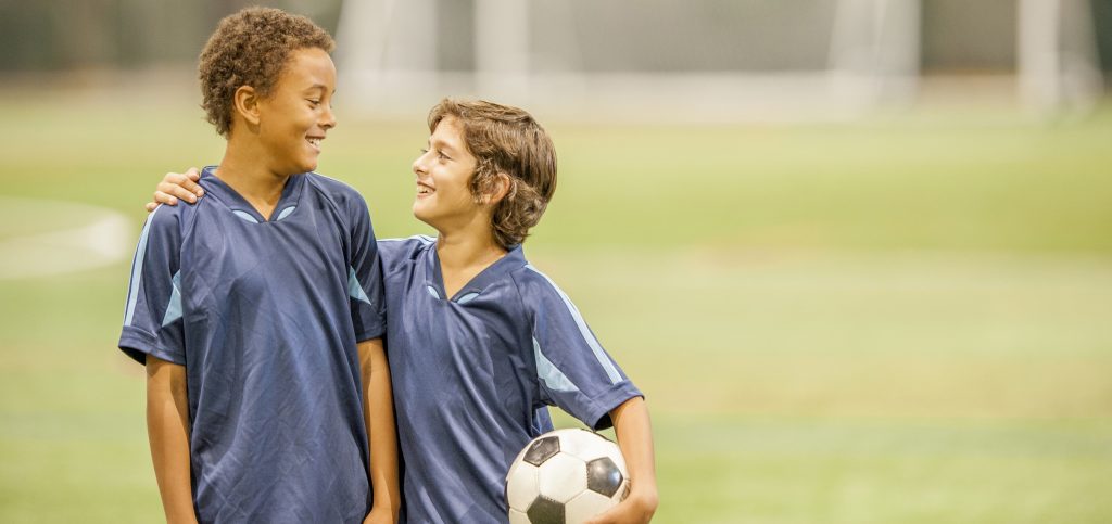 Two elementary age boys are standing together after the soccer game with a soccer ball.